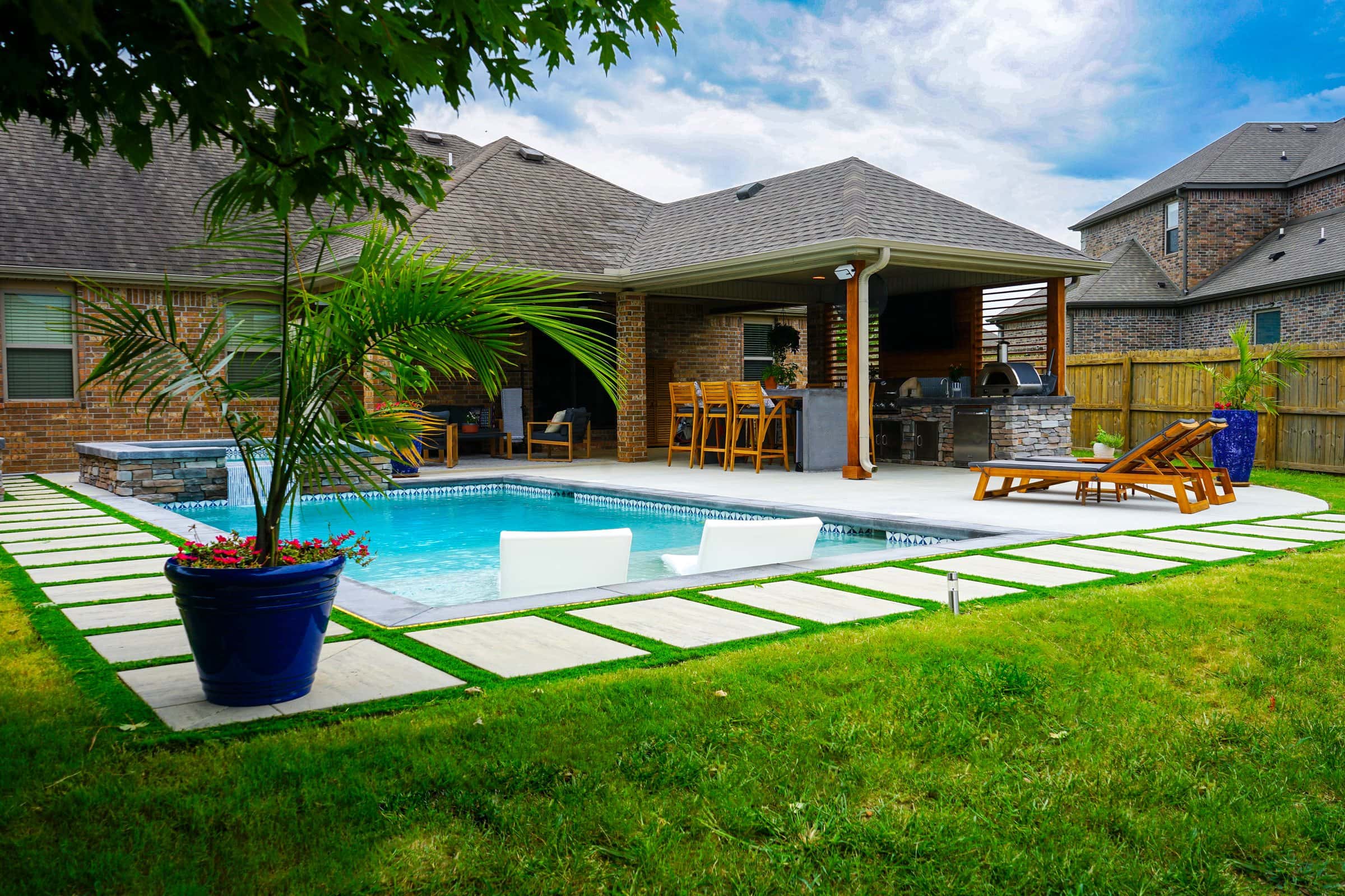 Backyard pool framed by turf-jointed pavers and green lawn with a potted palm and covered patio.