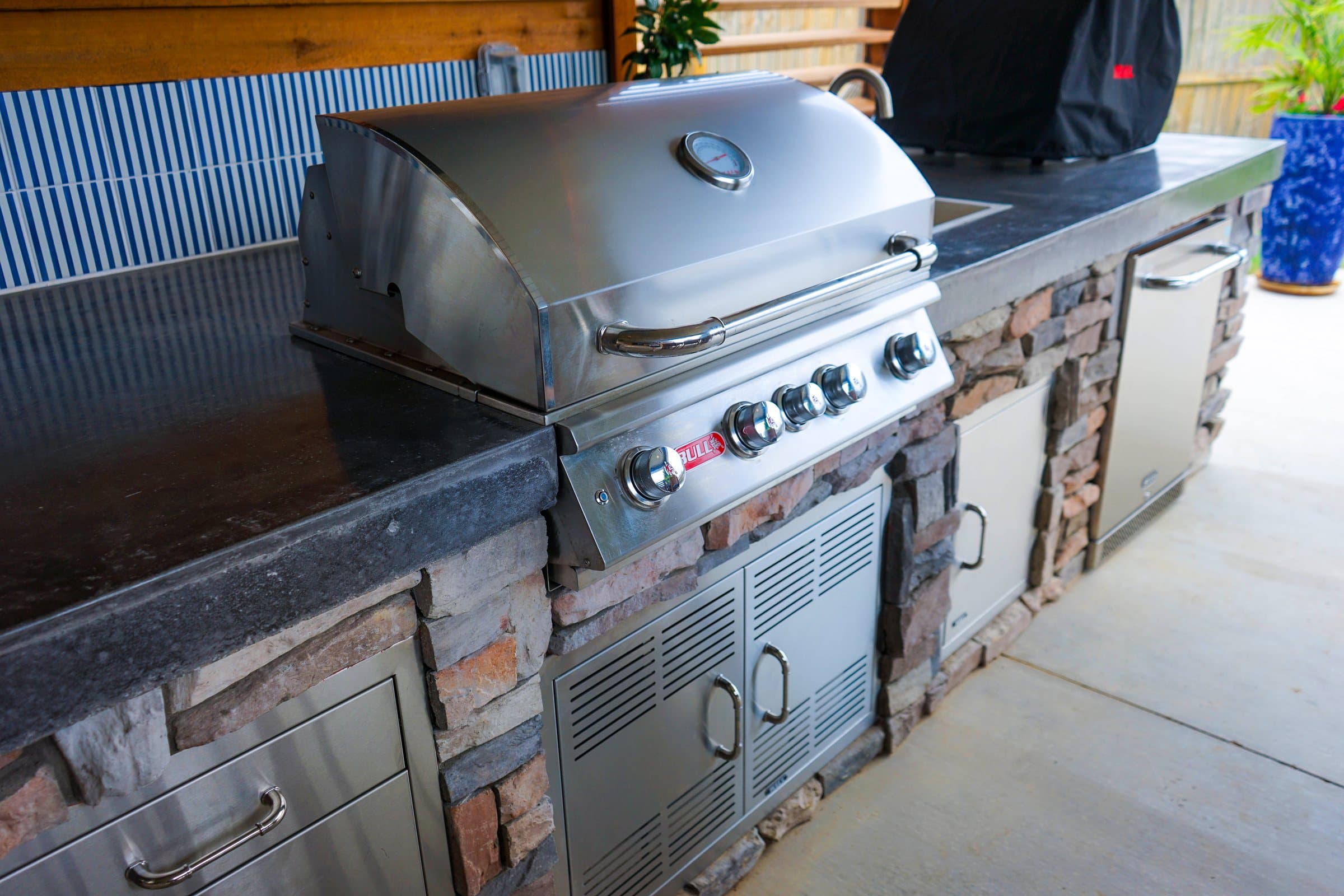 Stainless built-in grill and cabinet doors set into a stacked stone kitchen with dark counters.