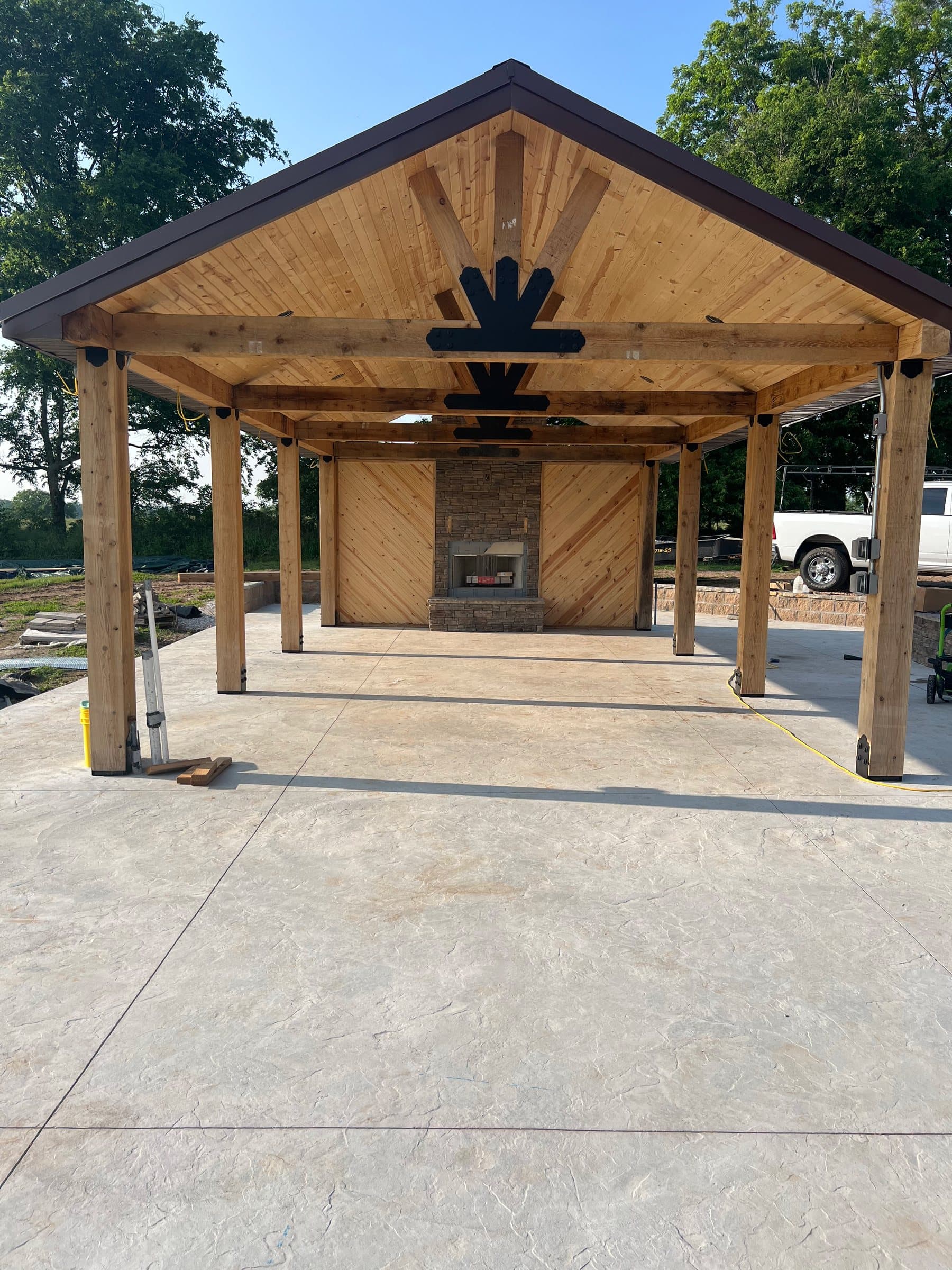 A cedar gable pavilion with a black sunburst bracket and stone fireplace on a stamped concrete slab.