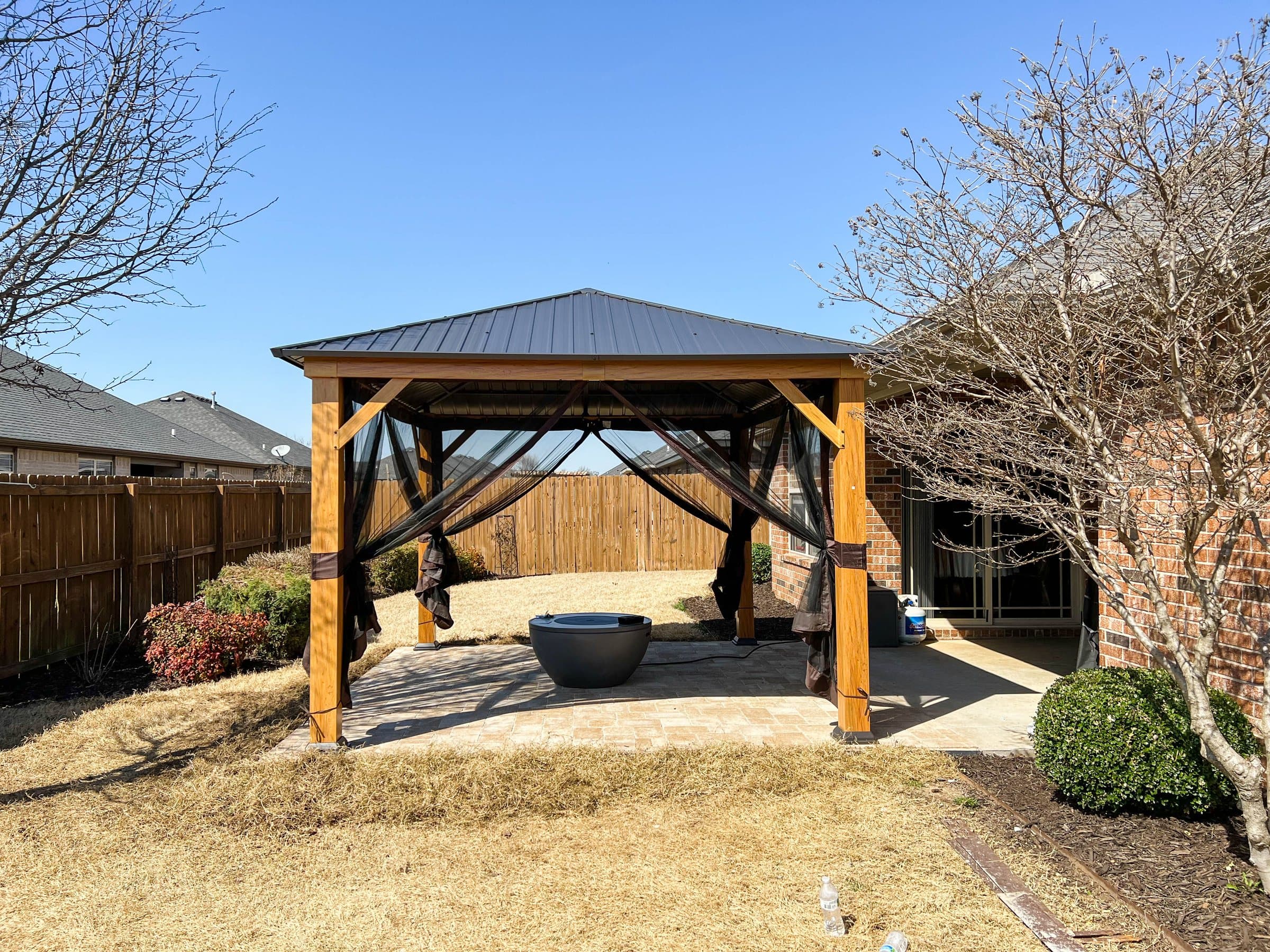 A cedar gazebo with a black metal roof and netting shelters a fire bowl on a patio.