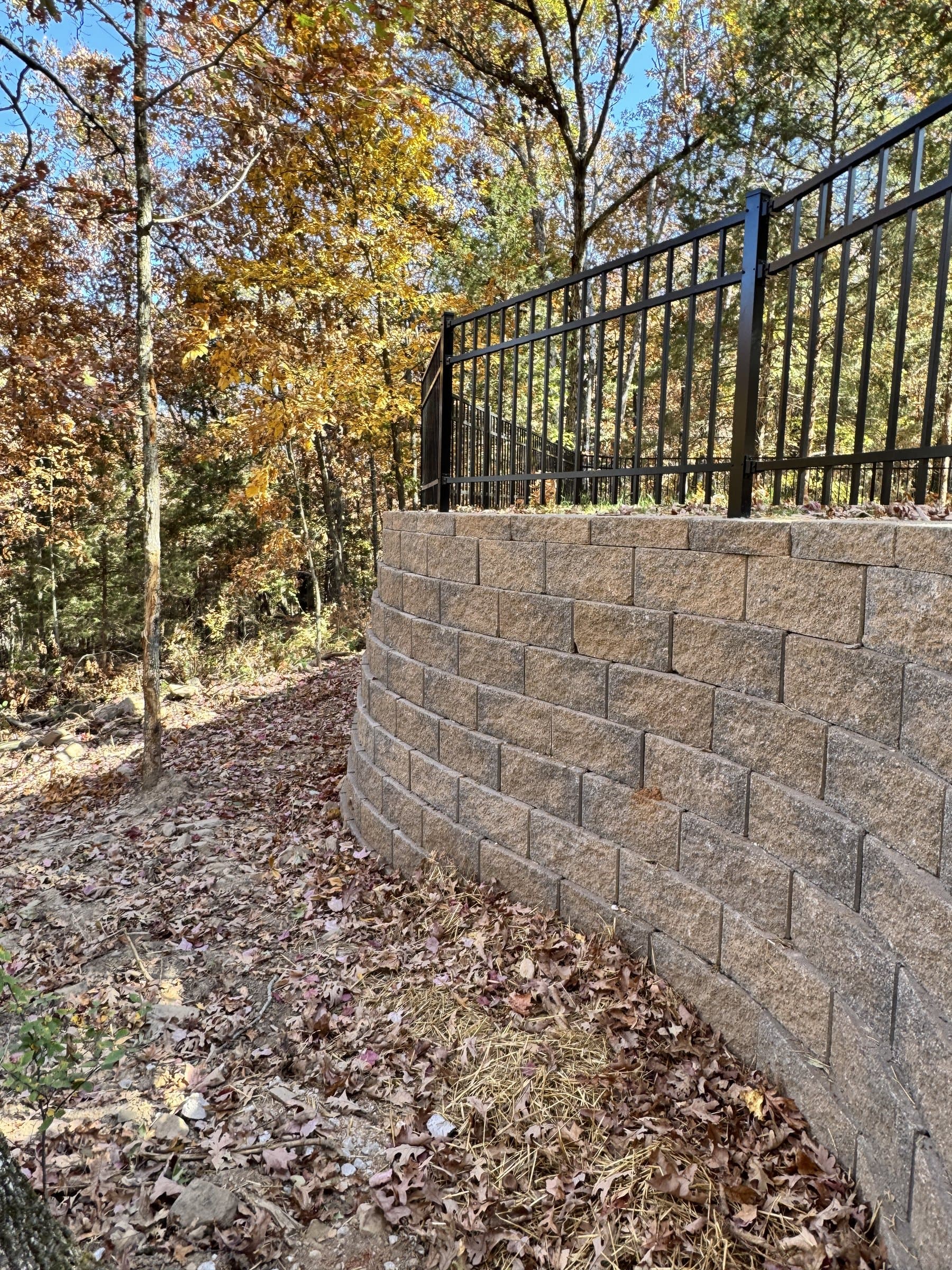 Curved tan block retaining wall with a black metal fence set among autumn trees.
