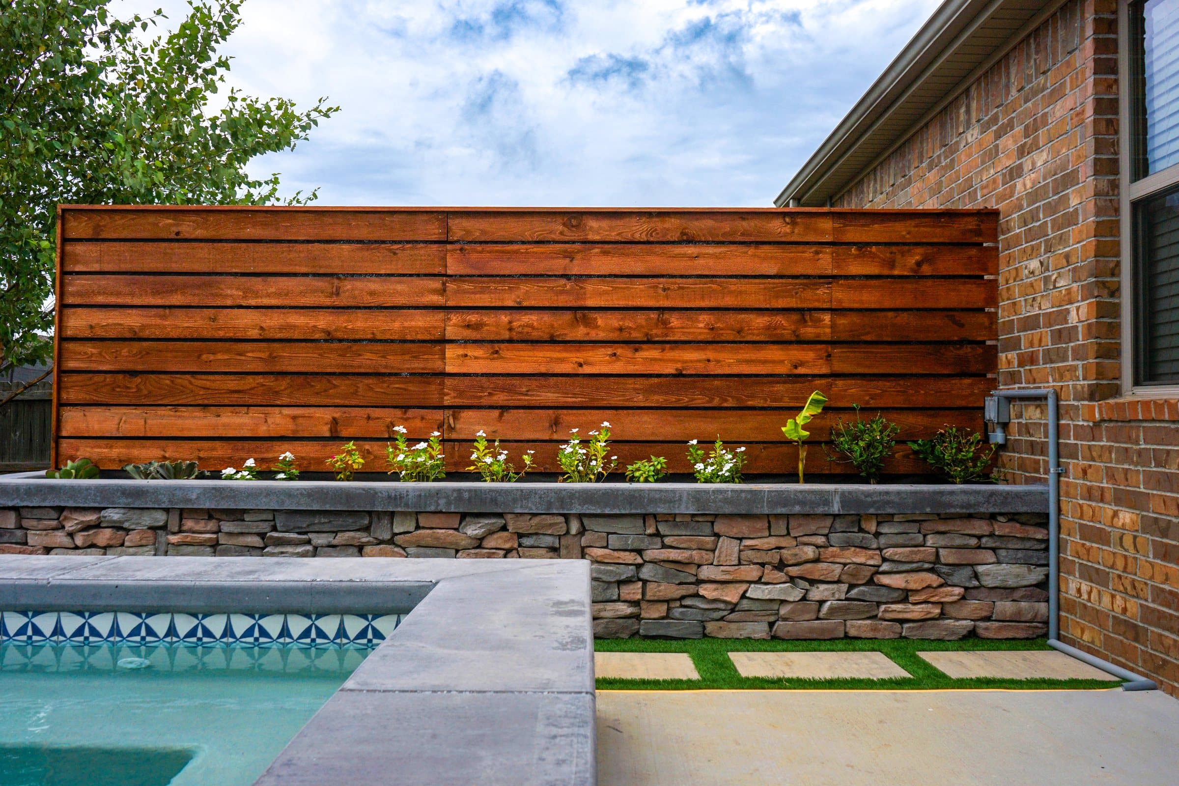 Horizontal cedar privacy screen rising above a stacked stone planter beside a spa.