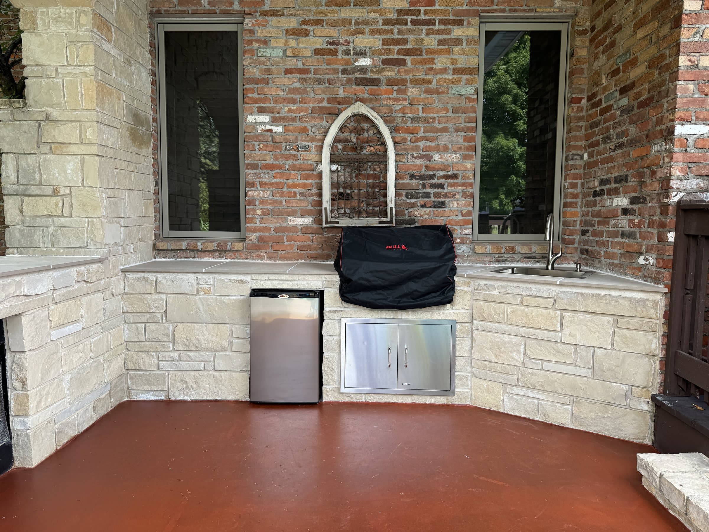 A covered outdoor kitchen with limestone counters, brick walls and a glossy red-brown stained concrete floor.