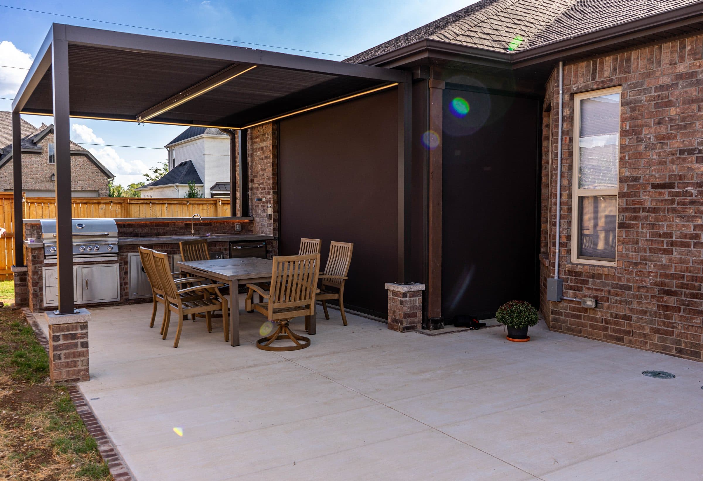 Dark louvered metal pergola over a concrete patio, with a brick outdoor kitchen and dining set beside a brick home.