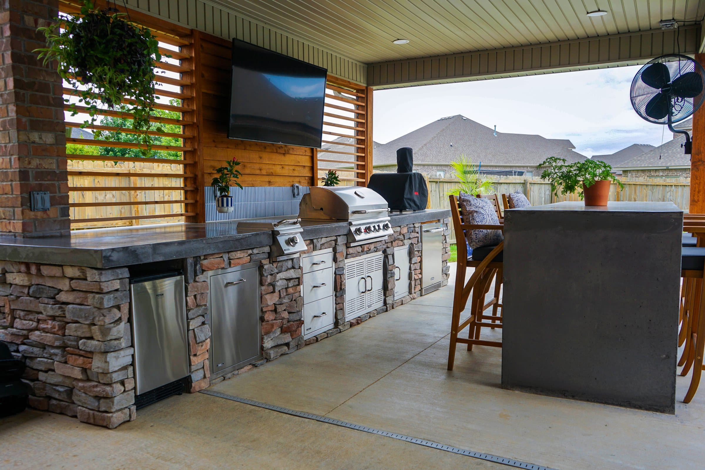 Covered outdoor kitchen with stacked stone base, grill, TV and a bar-height counter with stools.