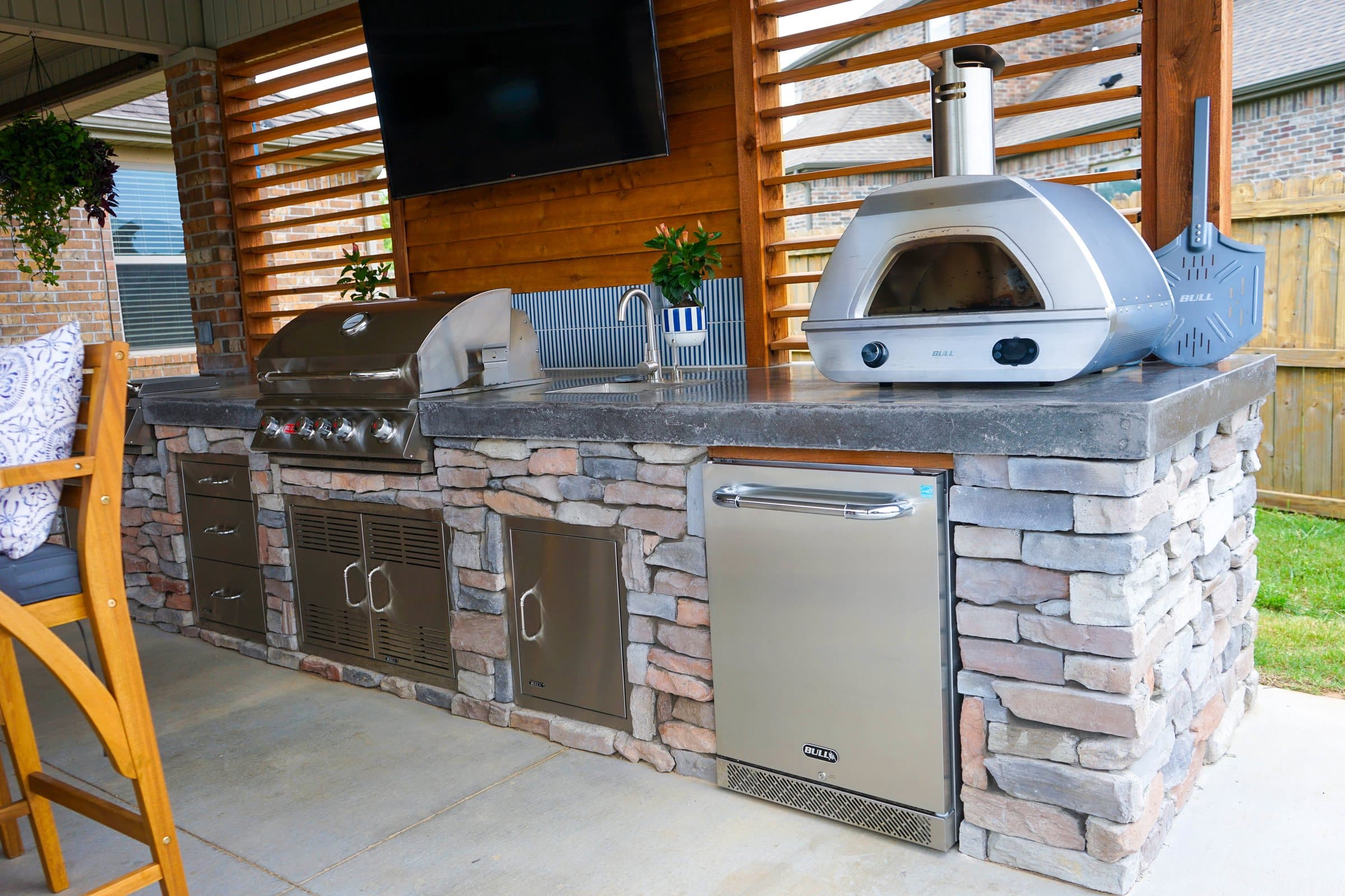 Stone outdoor kitchen with a pizza oven, grill and stainless fridge below a mounted TV.