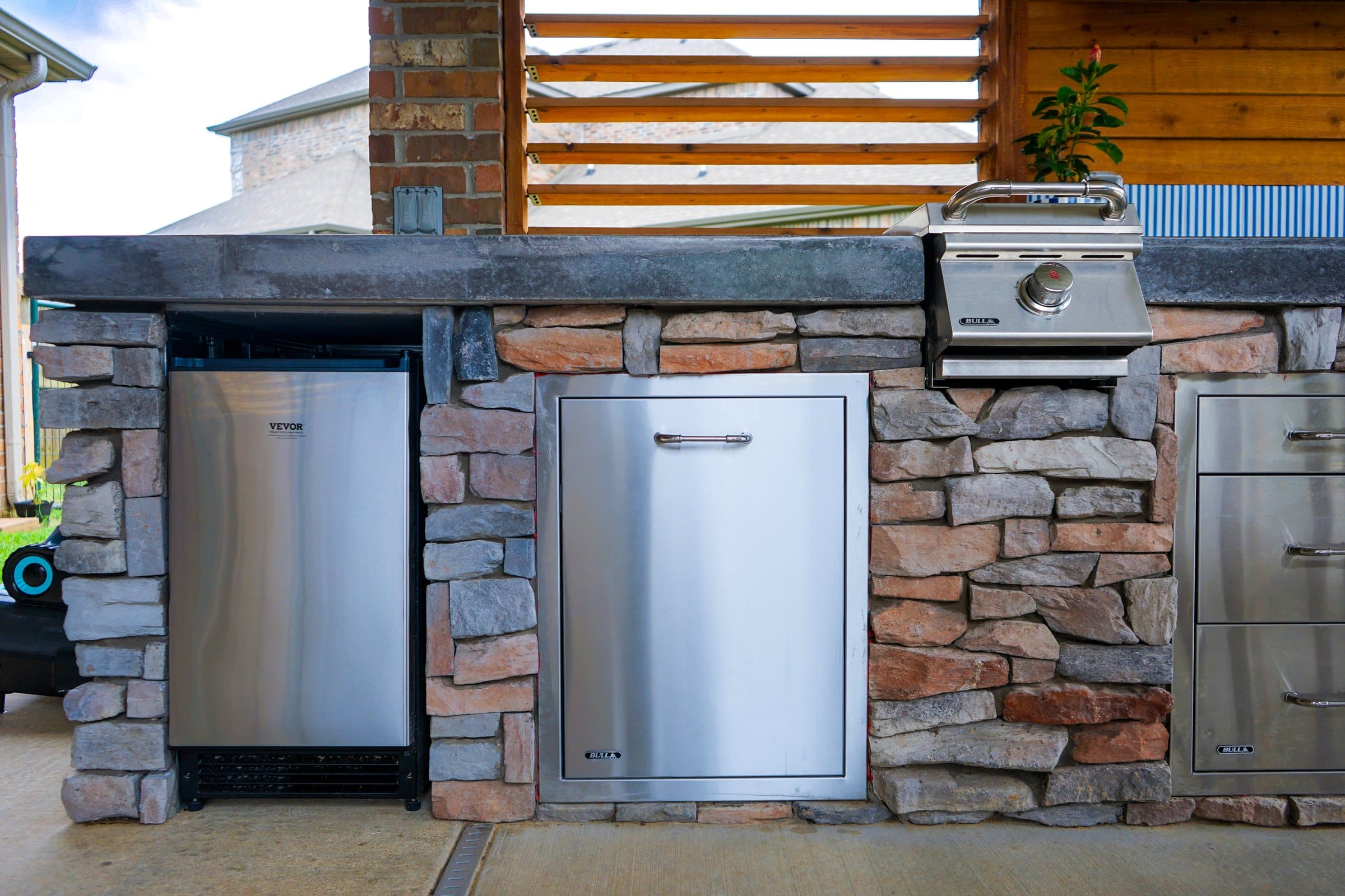 Stainless fridge, access doors and side burner built into a stacked stone outdoor kitchen.