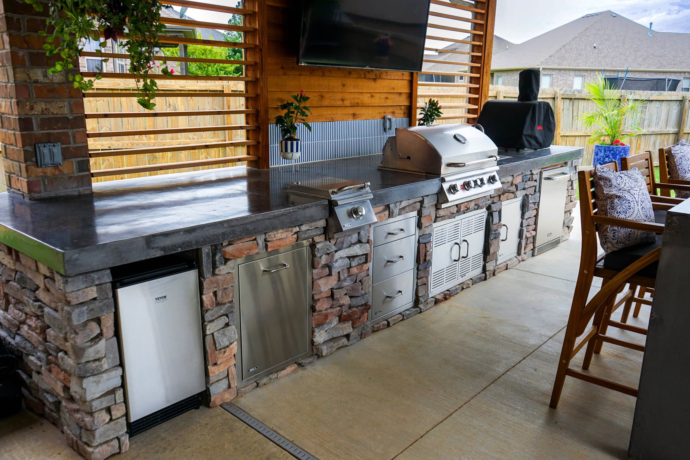 Outdoor kitchen with stacked stone base, stainless appliances and dark counters under a covered patio.