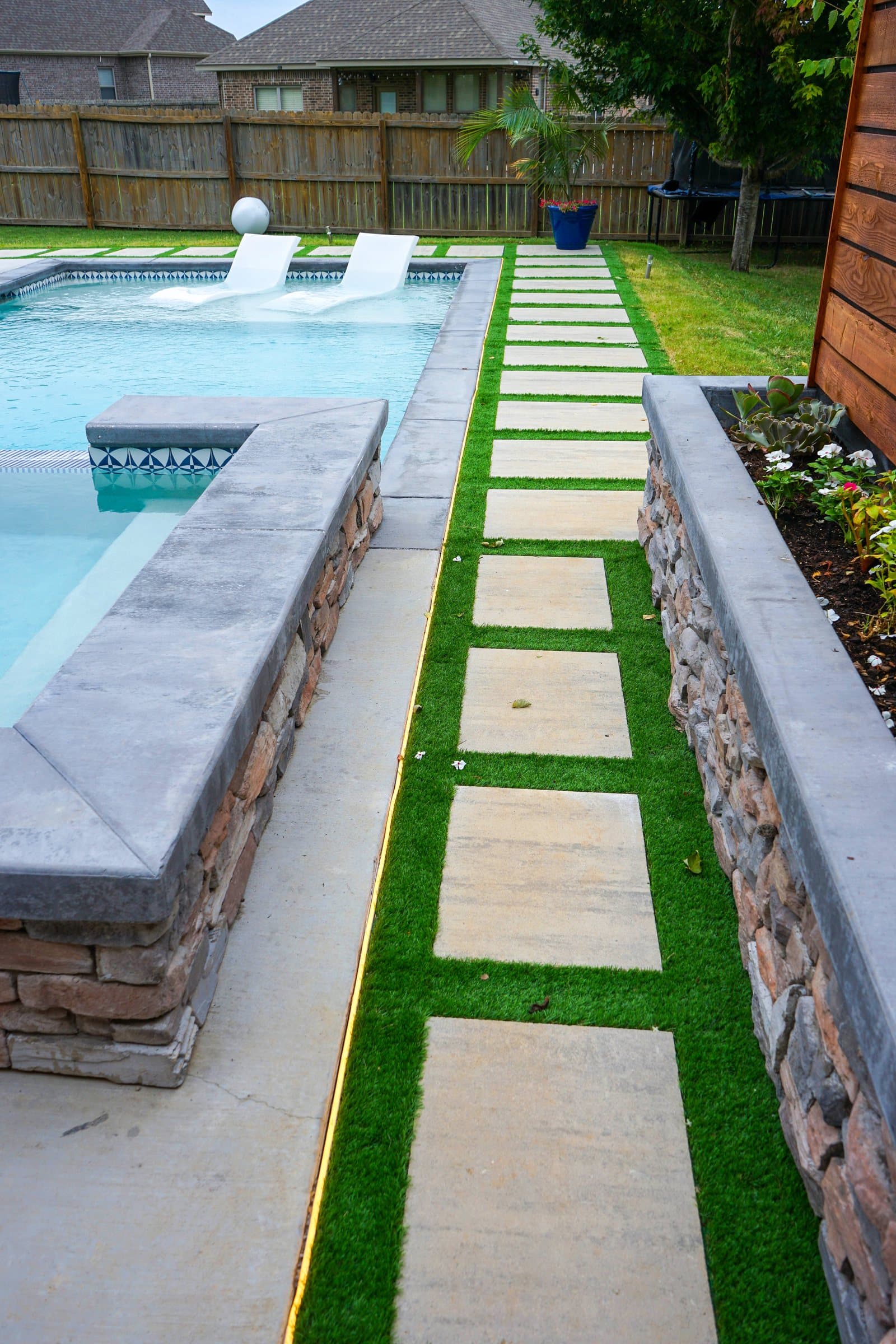 Large pavers set in artificial turf run between a pool's stone spa wall and a stone planter.