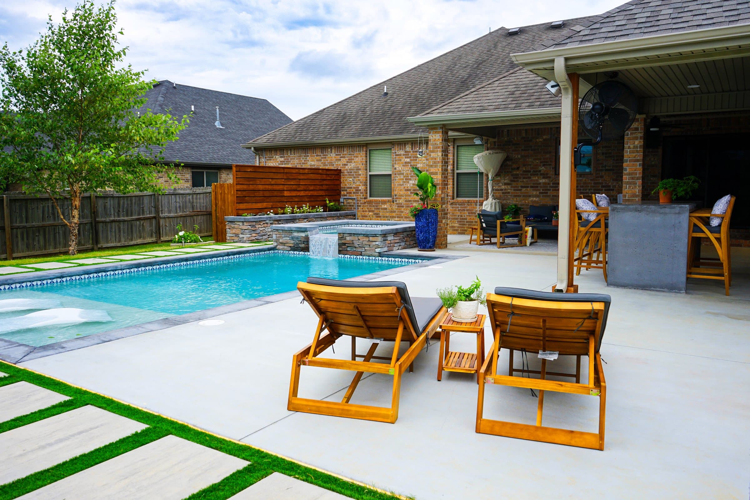 Teak lounge chairs on a concrete deck by a pool and spa with a covered bar behind.