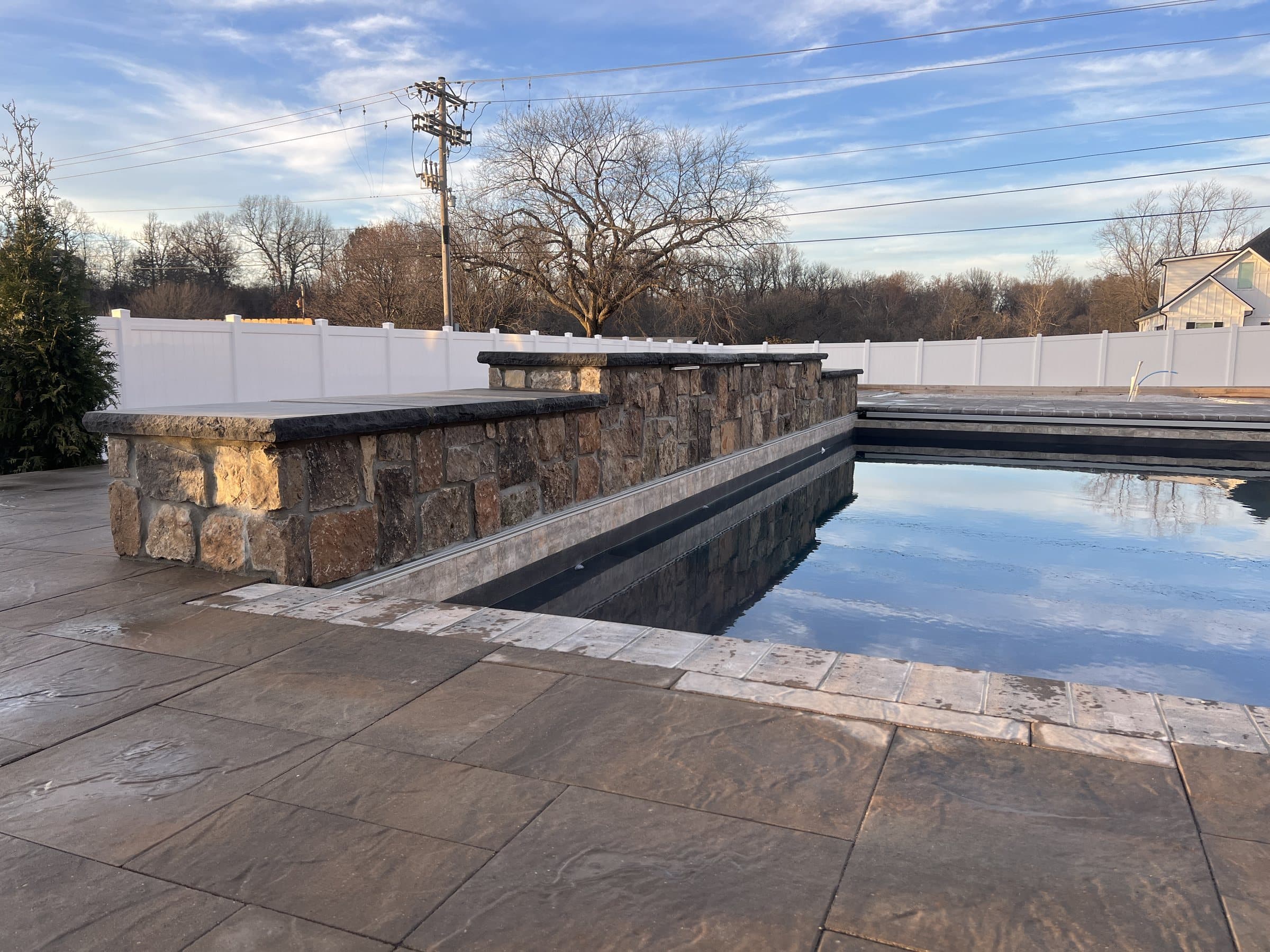 A pool reflects the sky beside a stone-veneer raised wall and large-format paver deck.