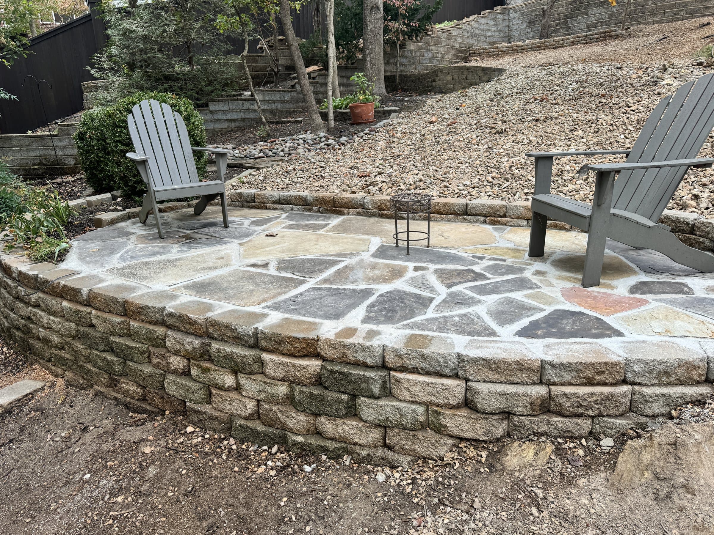 Raised round flagstone patio edged by a block wall with gray Adirondack chairs.