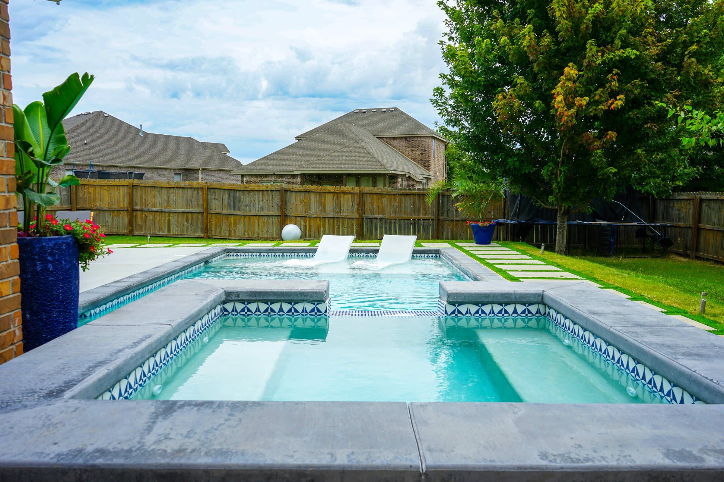 Raised spa with blue mosaic tile in front of a pool and tanning ledge by a wood fence.