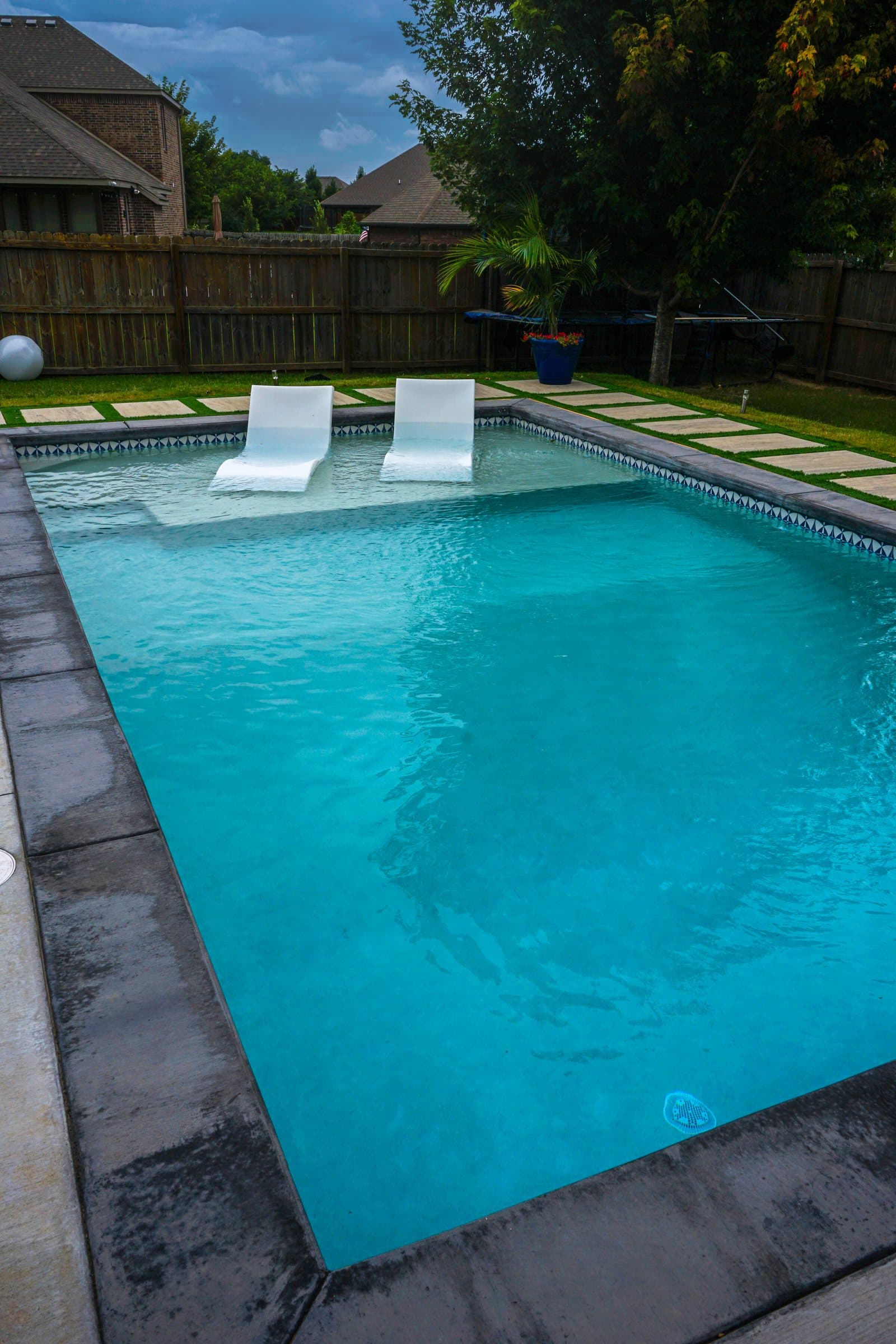 Long turquoise pool with dark coping and white loungers on the far tanning ledge.