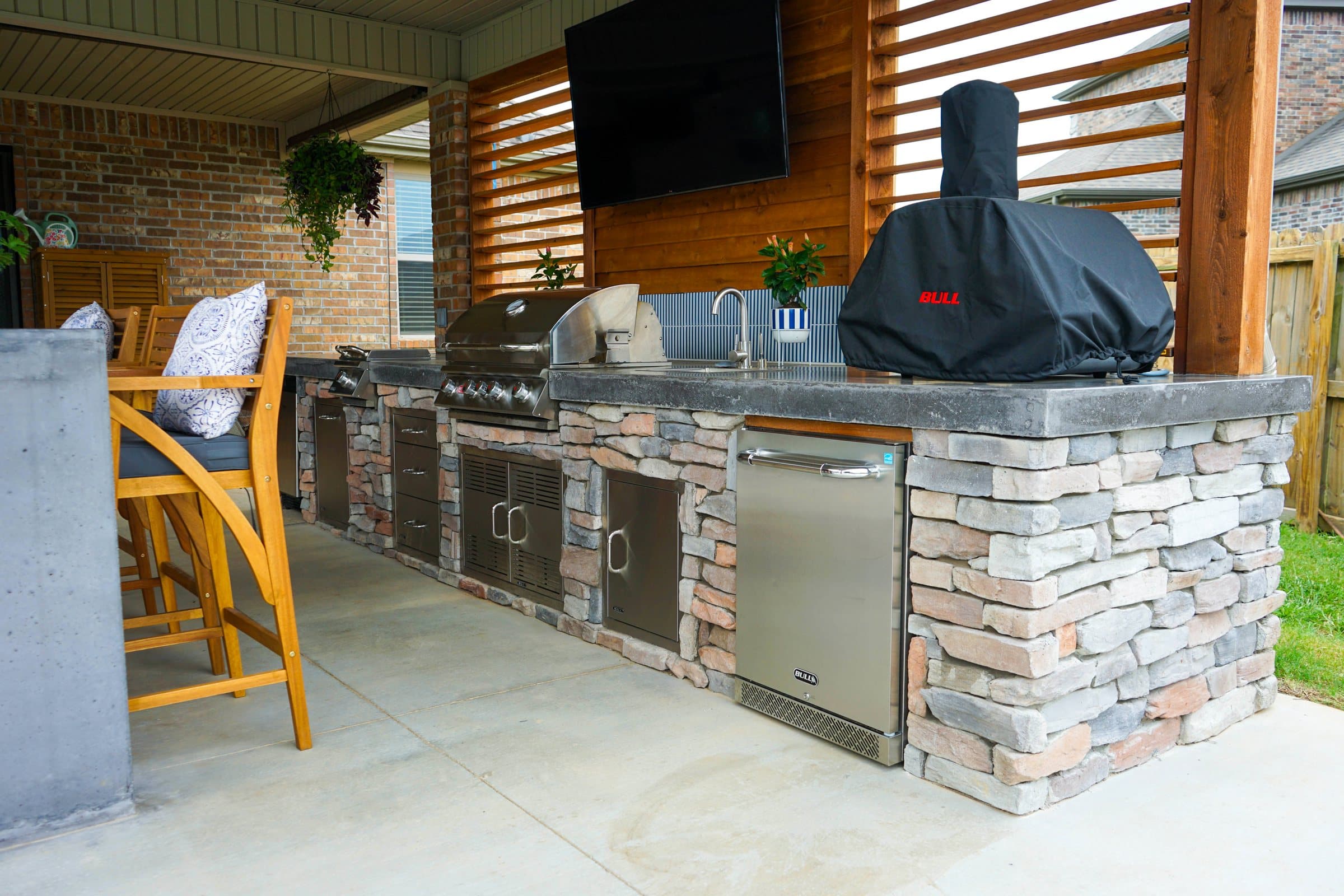 Stacked stone outdoor kitchen with stainless grill and fridge under a TV on a cedar slat wall.