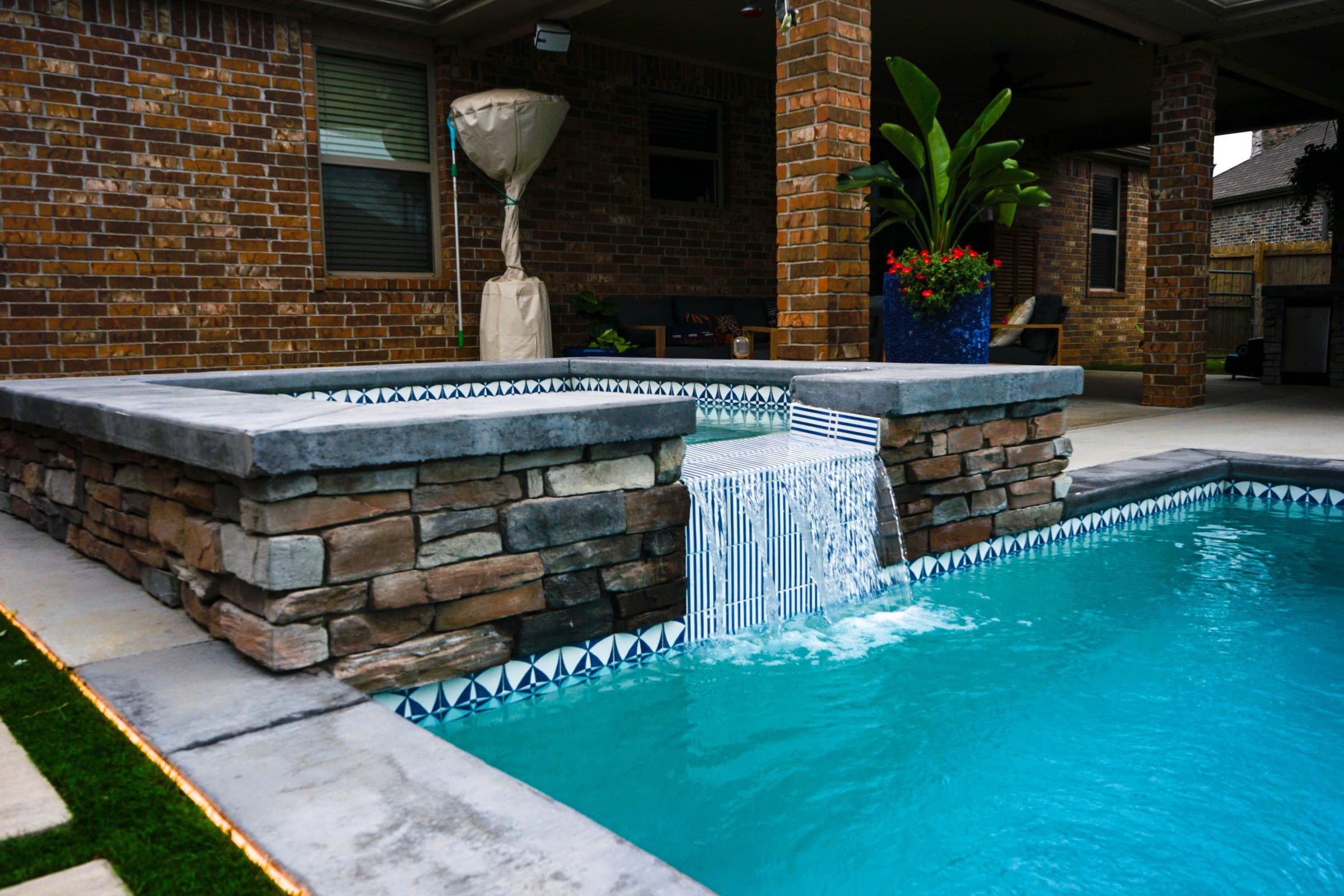 Raised stacked stone spa pouring a sheer waterfall into a turquoise pool with mosaic tile trim.
