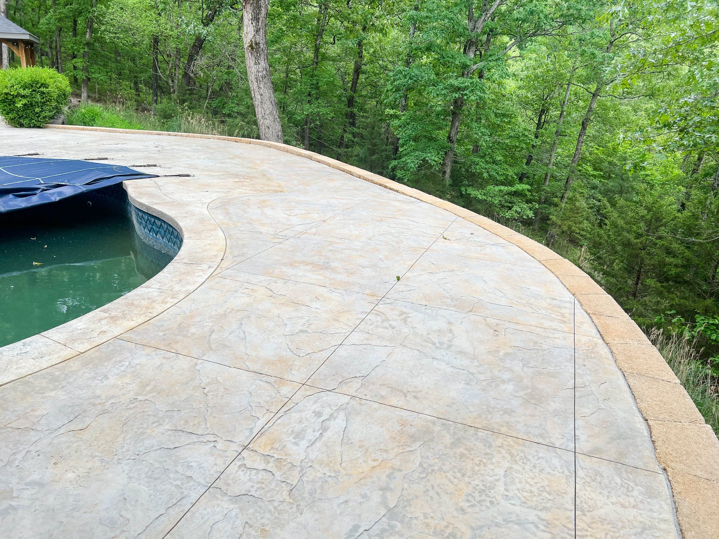 A tan stamped concrete pool deck curves around a covered pool against a wooded hillside.