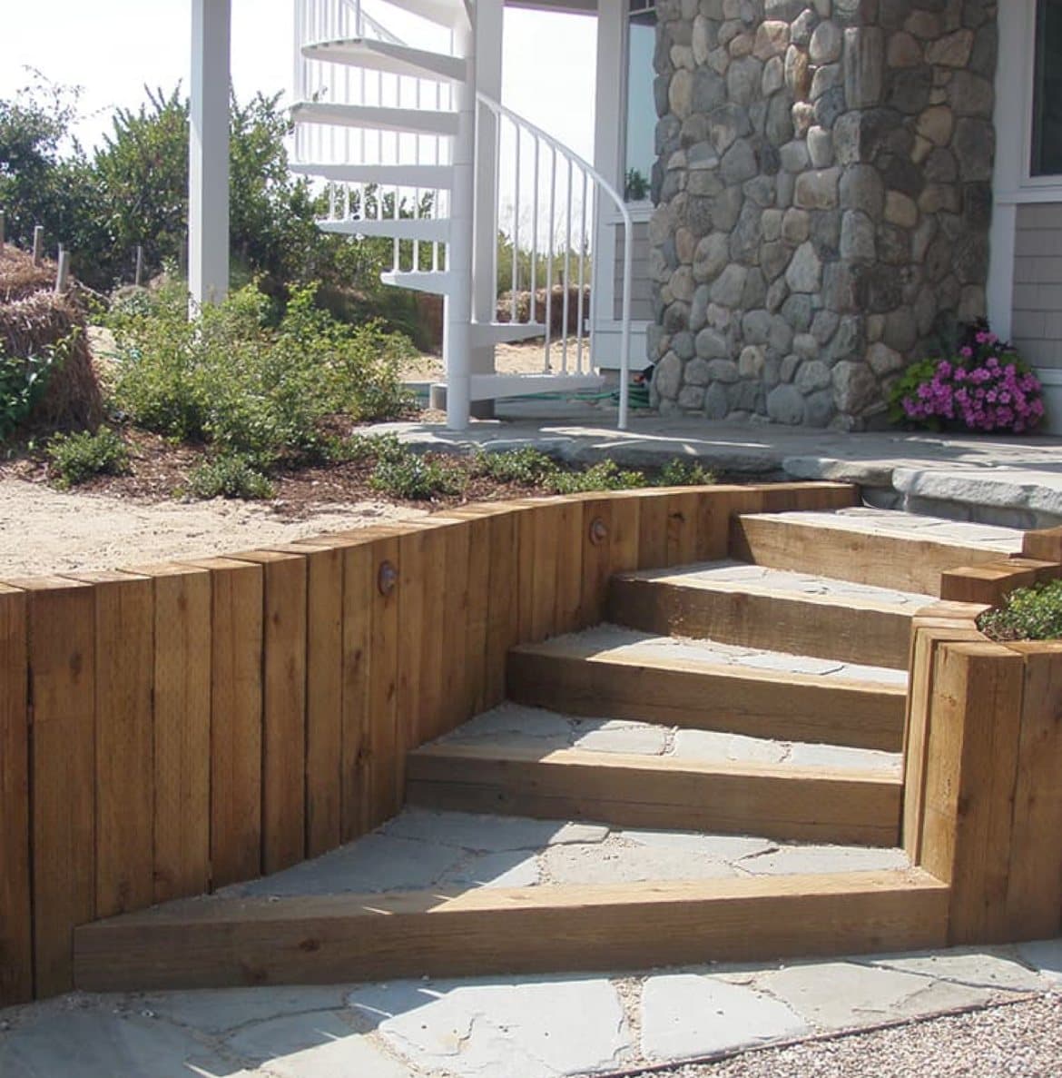 Wide timber steps with a flagstone landing climb toward a white spiral staircase and stone column.